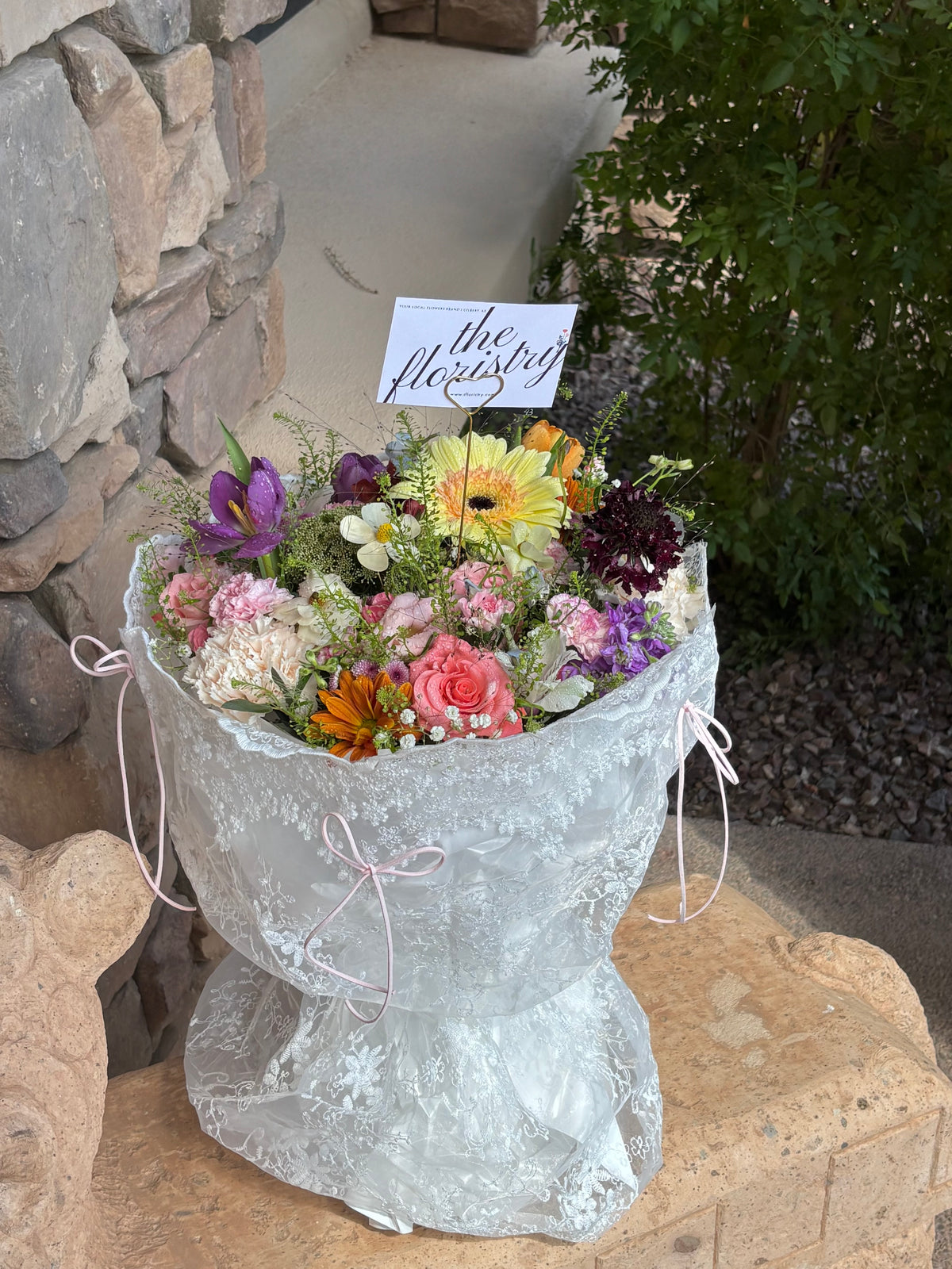A large, vibrant bouquet of assorted flowers wrapped in white lace paper with pink ribbon ties. Same-day flowers delivery in Gilbert Arizona.