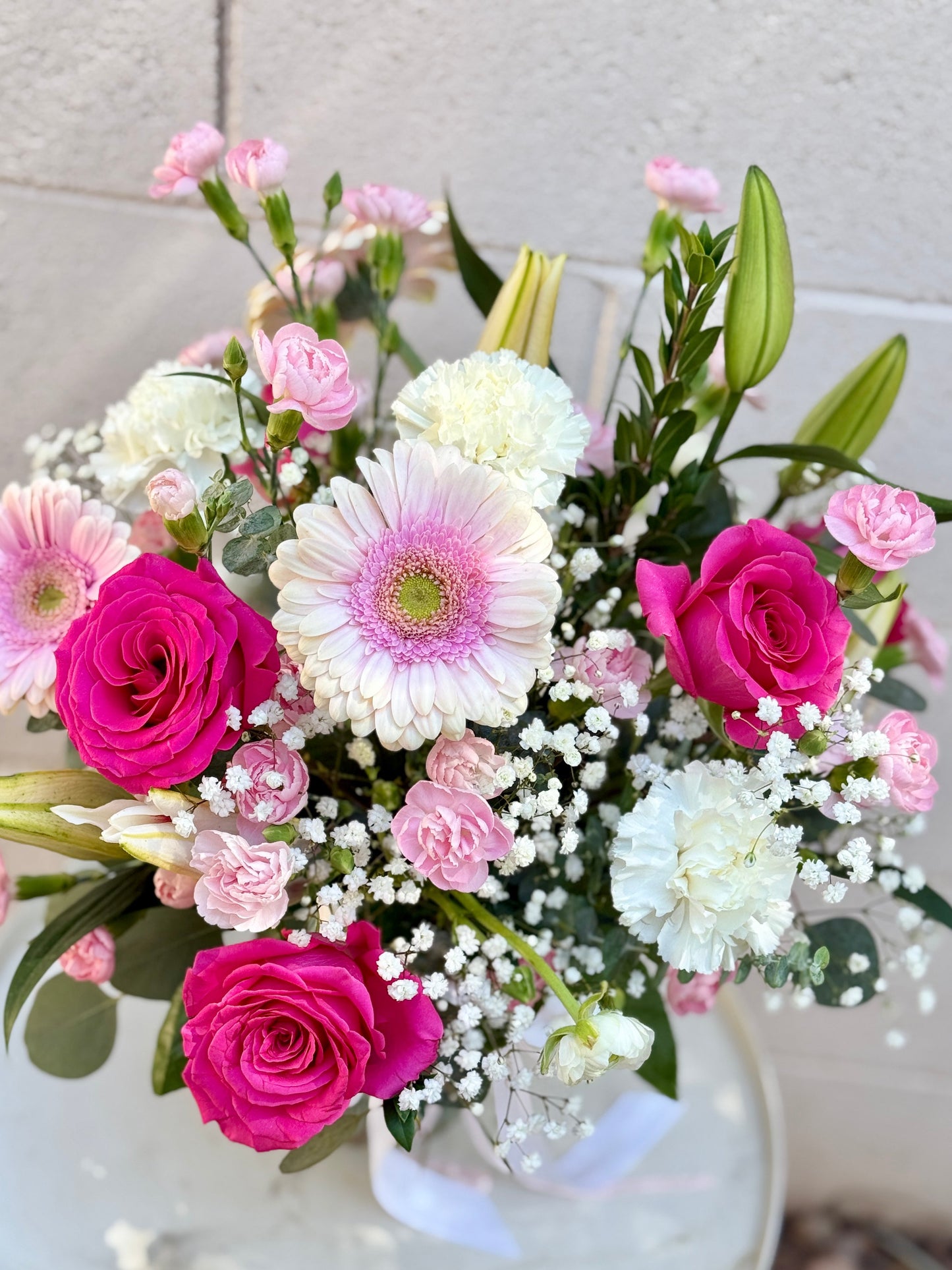 A vibrant flower bouquet featuring bright pink roses, white and pink gerbera daisies, pink carnations, baby's breath, and green lily buds, arranged in a white vase