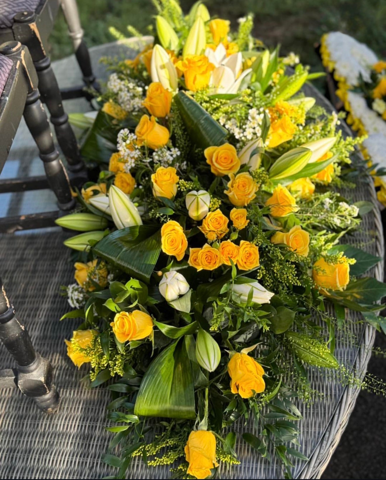 A large yellow and white floral arrangement with roses, lilies, and green foliage, displayed on a woven chair outdoors.