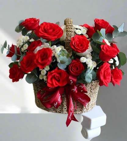A basket arrangement of red roses with green leaves and white flowers, adorned with a red ribbon.
