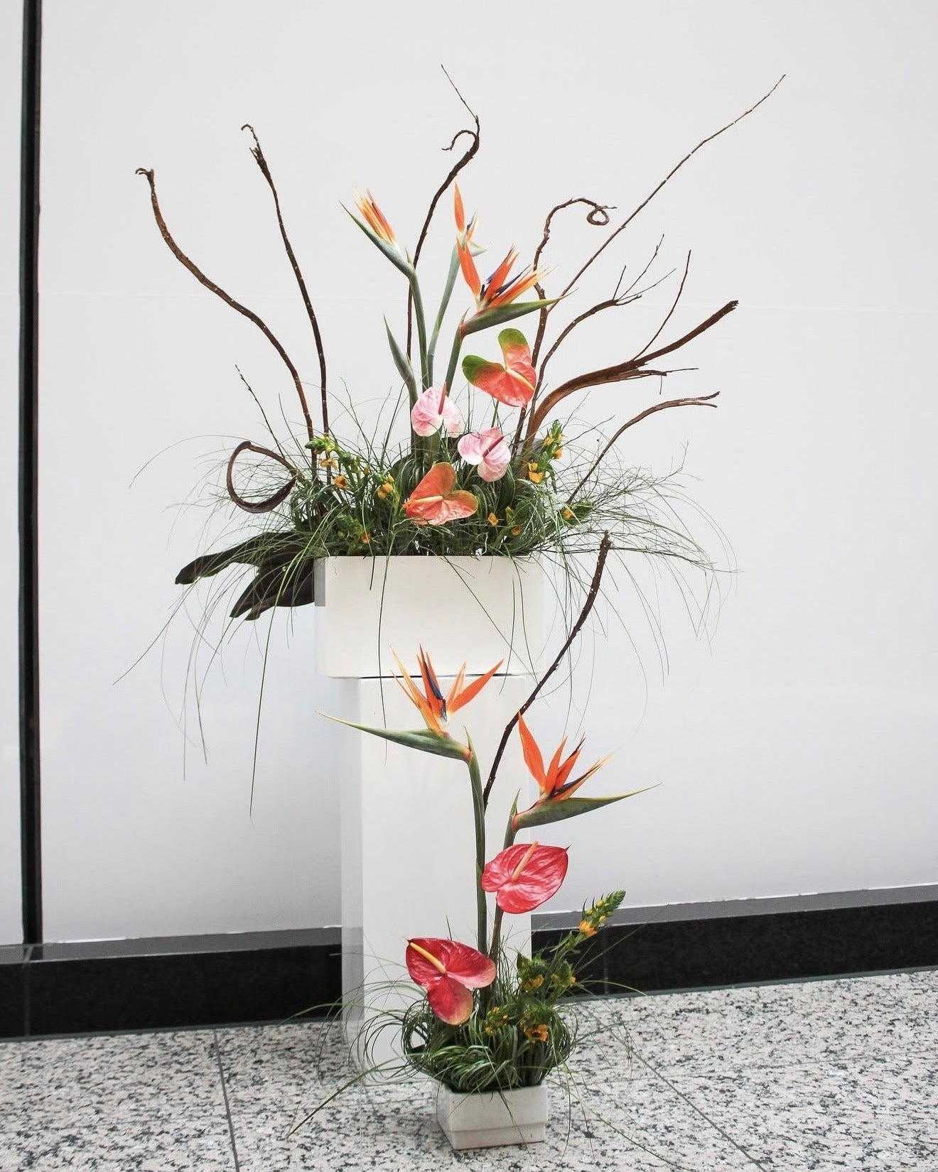 A decorative floral arrangement featuring orange bird of paradise flowers, pink and red anthuriums, green foliage, and brown branches, displayed in a white pedestal planter against a white wall.