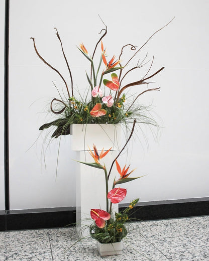 A decorative floral arrangement featuring orange bird of paradise flowers, pink and red anthuriums, green foliage, and brown branches, displayed in a white pedestal planter against a white wall.