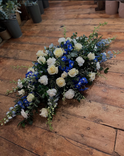 A floral arrangement featuring white and cream roses, blue delphiniums, and green foliage, displayed on a rustic wooden floor.