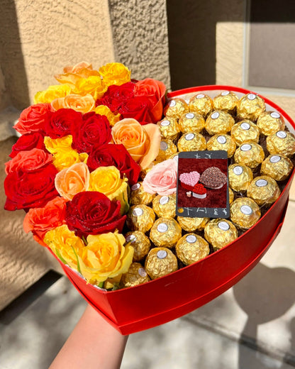 A heart-shaped box containing an arrangement of red and yellow roses along with Ferrero Rocher chocolates.
