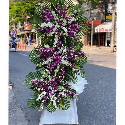 Luxurious standing funeral tribute featuring vibrant purple orchids, palm leaves, and lavender wrapping.