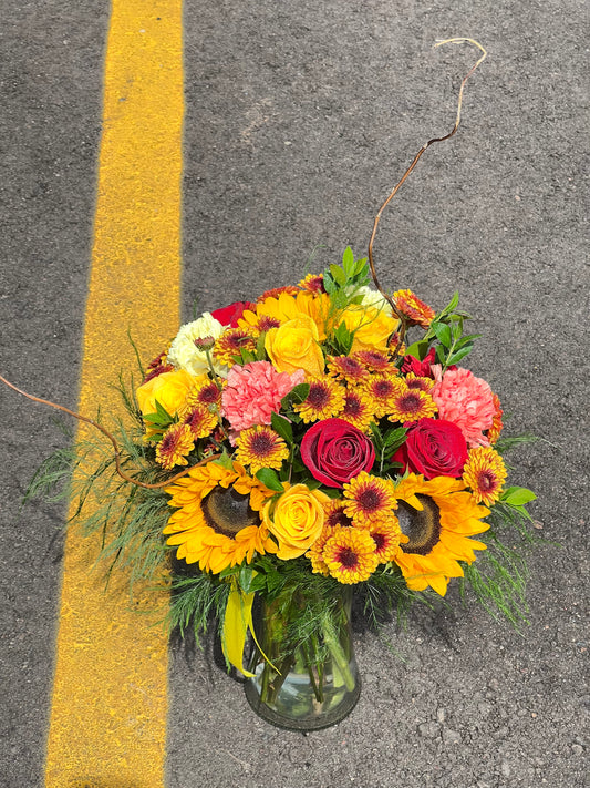 A vibrant Harvest Bouquet with a variety of flowers including roses and sunflowers, arranged in a clear glass vase, yellow, red, orange and warm tone.