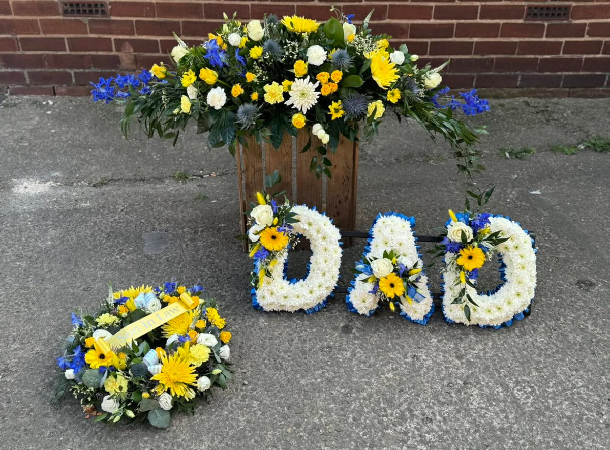 A floral memorial arrangement on a concrete surface against a brick wall, featuring a large flower arrangement in a wooden crate behind it, a round matching flower wreath in the foreground, and three white flower letters spelling DAD with blue and yellow floral accents.