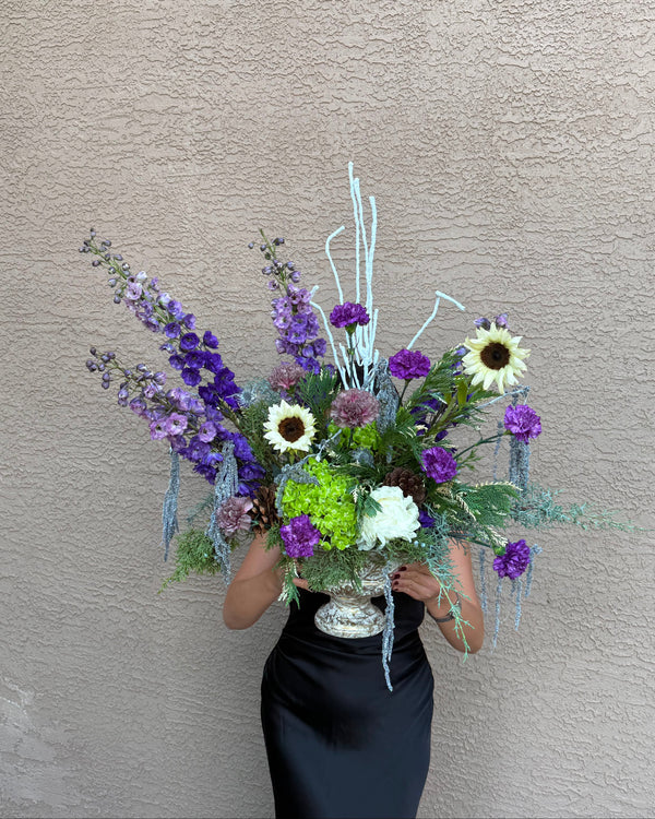 a large, elaborate floral arrangement featuring purple delphiniums, white sunflowers, green foliage, and light blue dried stems in a silver urn