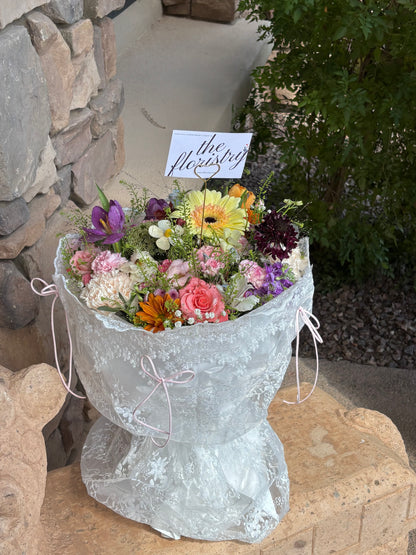 A large, vibrant bouquet of assorted flowers wrapped in white lace paper with pink ribbon ties. Same-day flowers delivery in Gilbert Arizona.