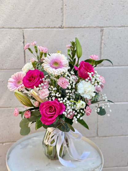 A vibrant floral bouquet featuring pink and white roses, daisies, baby's breath, and green foliage, arranged in a clear glass vase with a white ribbon, set on a marble-topped table against a light gray brick wall.