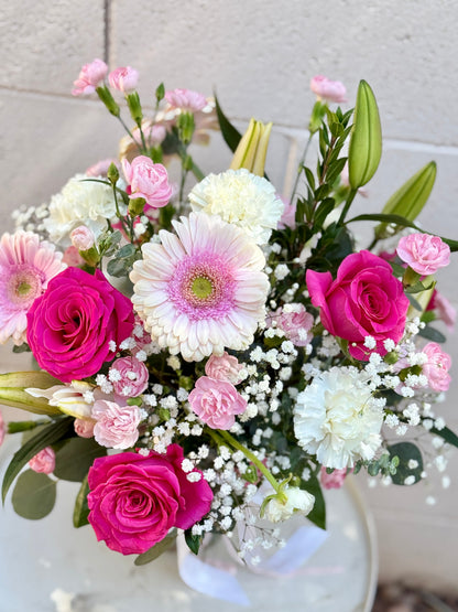A vibrant flower bouquet featuring bright pink roses, white and pink gerbera daisies, pink carnations, baby's breath, and green lily buds, arranged in a white vase 
