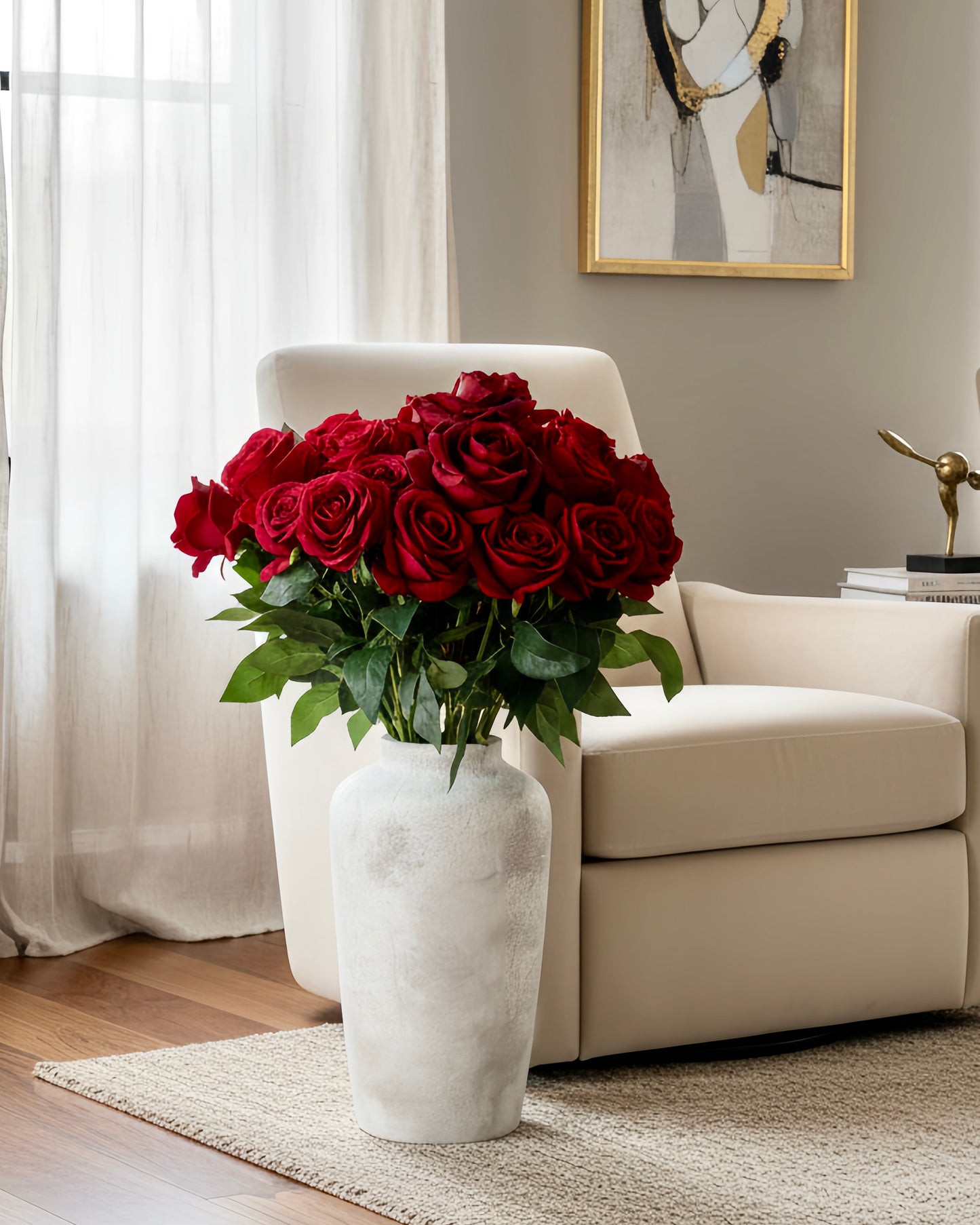 A large bouquet of deep red roses with green leaves arranged in a tall textured white vase, placed in front of a cream-colored armchair in a softly lit living room.