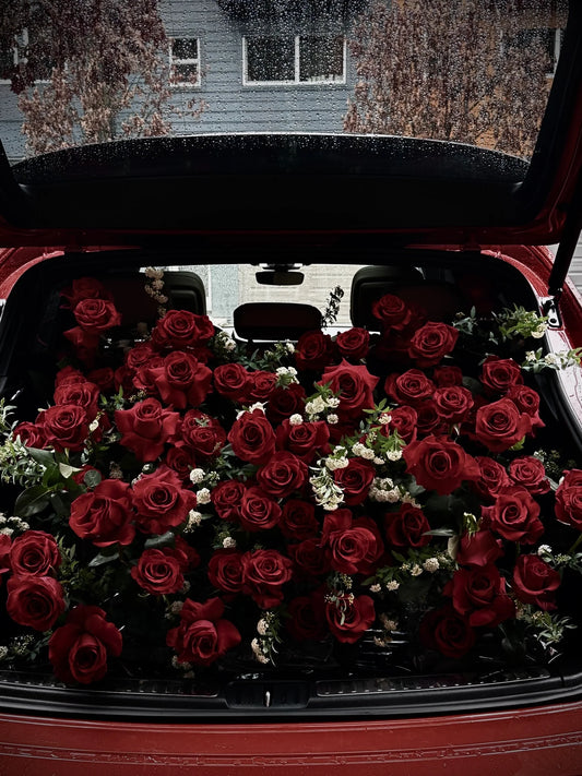  Deep red roses with green leaves and small white filler flowers arranged in the open trunk of a luxury red car.