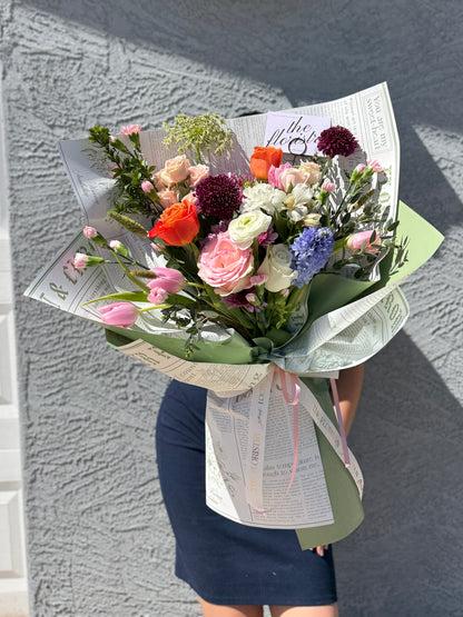 a large, vibrant bouquet of mixed flowers wrapped in newspaper-patterned paper and a sage green ribbon