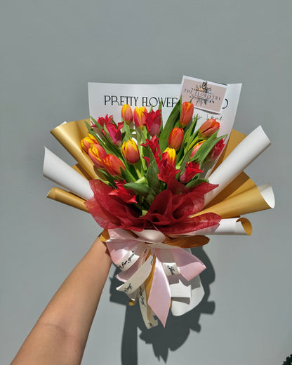 Bouquet of red and yellow tulips wrapped in gold and white paper, held against a gray background.