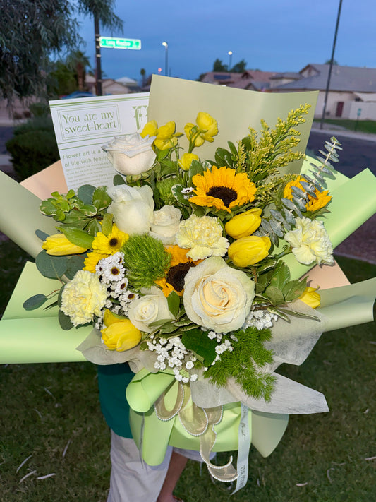 A vibrant flower bouquet held outdoors at dusk, featuring yellow tulips, white and yellow roses, sunflowers, green foliage, and white baby's breath, wrapped in pale green paper with a matching ribbon and a printed card.