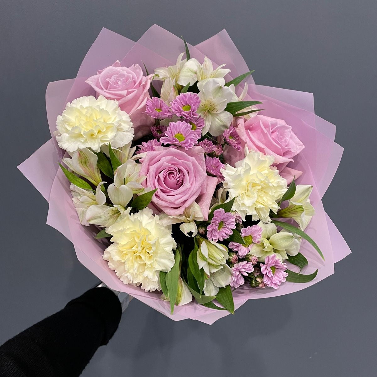 A bouquet of flowers featuring carnations and roses in shades of pink, white, and green, wrapped in pink paper.