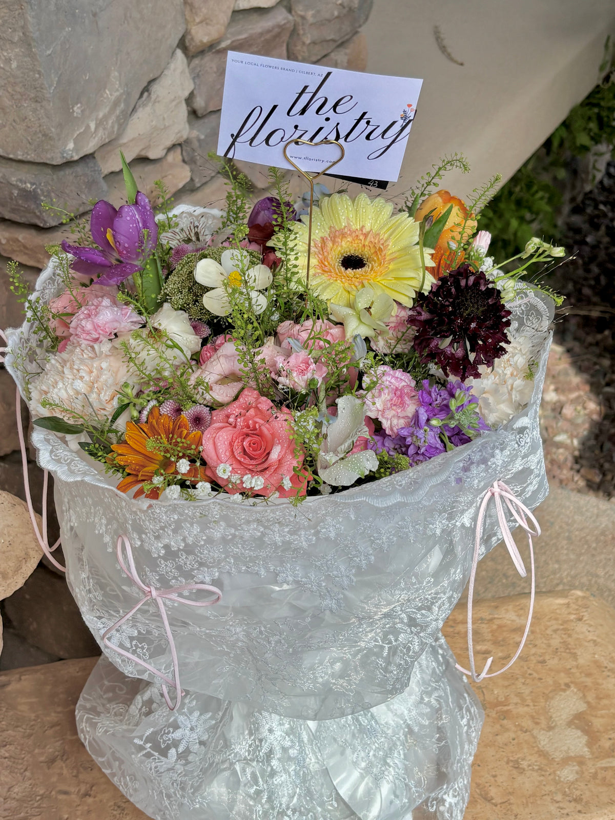 A vibrant mixed flower bouquet wrapped in textured lace style. The bouquet contains a variety of colorful flowers including pink roses, gerbera daisies, tulips. Same-day flowers delivery in Gilbert Arizona.