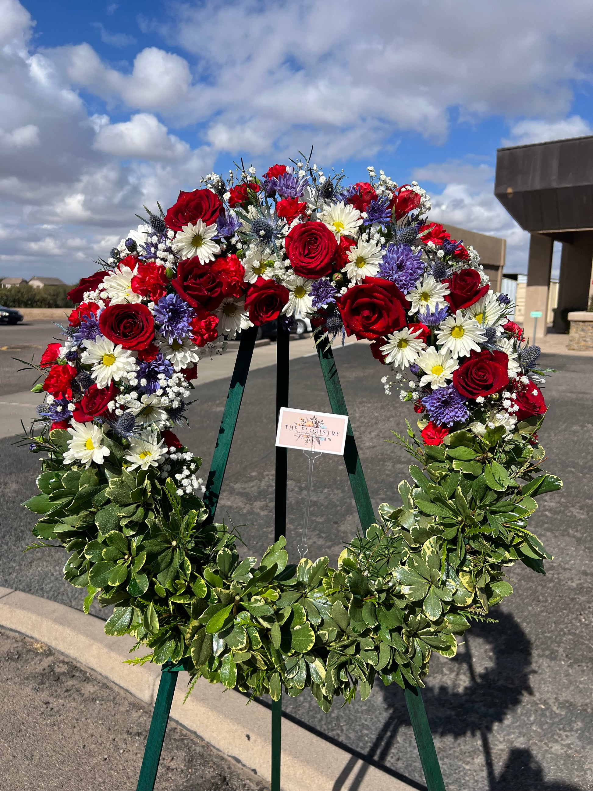 A round floral wreath with a patriotic color scheme of red, white, and blue, featuring various flowers and a card attached to it.