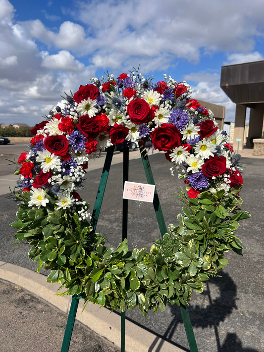 A round floral wreath with a patriotic color scheme of red, white, and blue, featuring various flowers and a card attached to it.