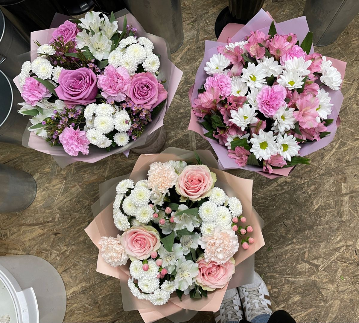 A bouquet of flowers featuring carnations and roses in shades of pink, white, and green, wrapped in pink paper.