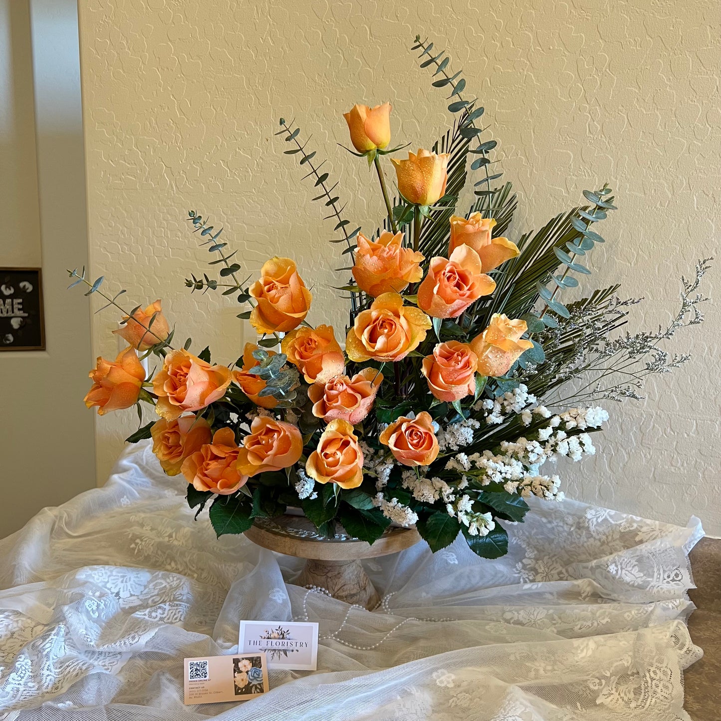 A floral arrangement featuring orange roses, white flowers, and green foliage in a basket, presented on a table with a white cloth.